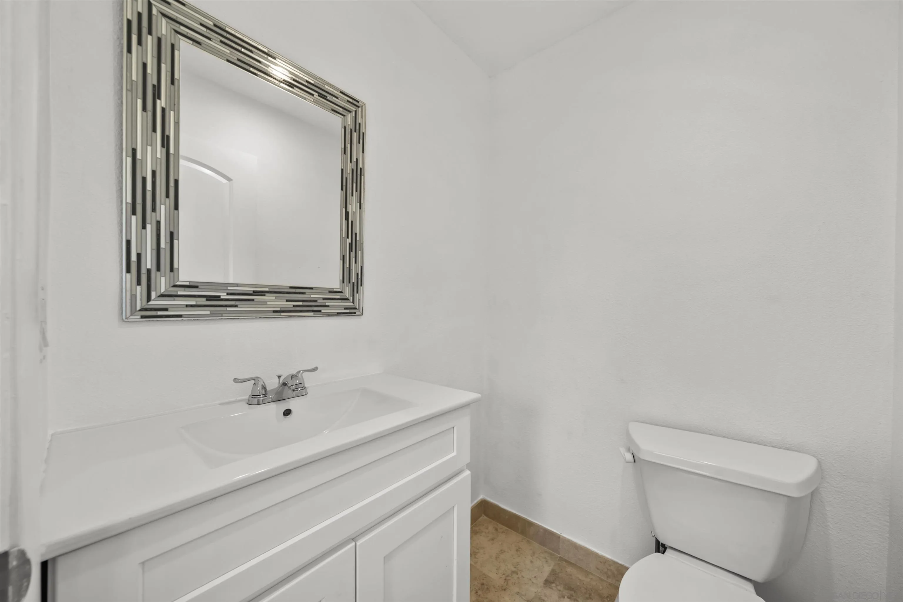 Bathroom with a white vanity, geometric-framed mirror, toilet, and tan tile floor.