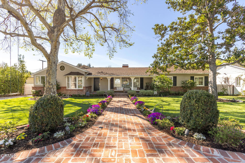 Single-story home framed by mature trees, with a brick walkway and colorful flower beds.