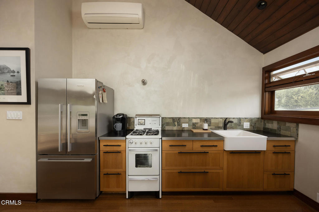 Kitchen with warm wood cabinetry, stainless steel refrigerator, white range, and farmhouse sink beneath a vaulted ceiling.
