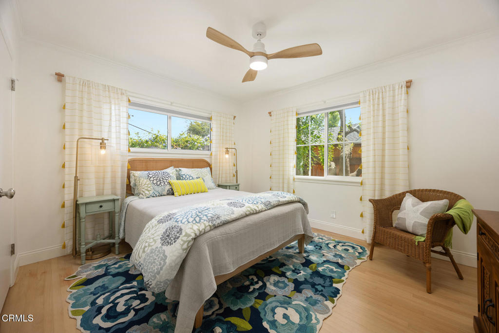 Bedroom with floral bedding and rug, two curtained windows, and a wicker chair.