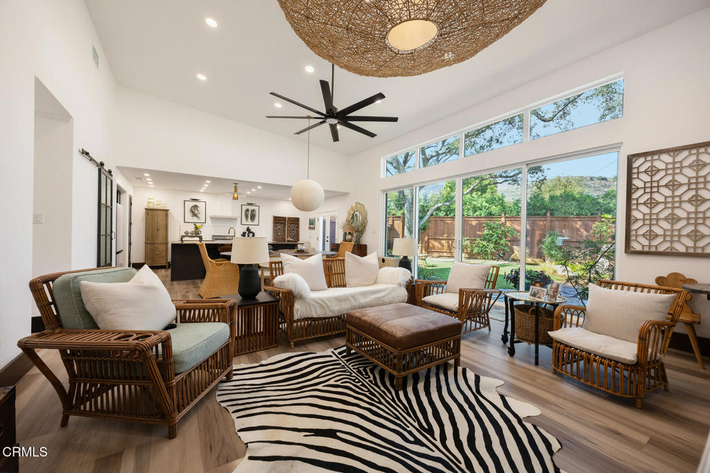 Living room with rattan seating, zebra-pattern rug, and tall windows overlooking a leafy yard.