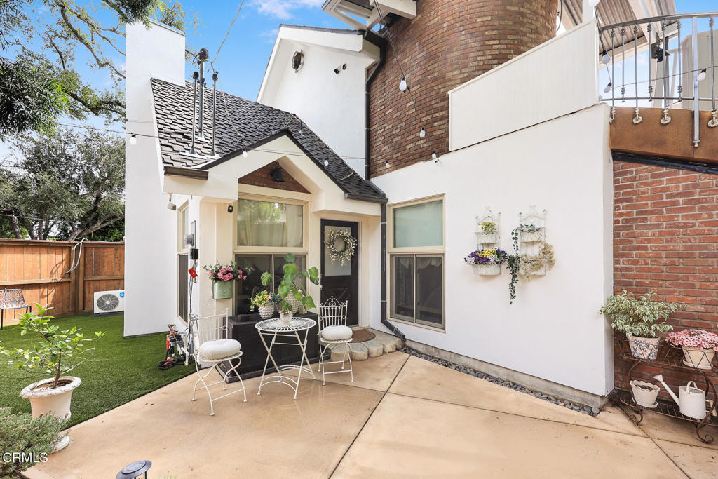 Rear courtyard with a concrete patio, café seating, potted flowers, and a fenced artificial lawn.