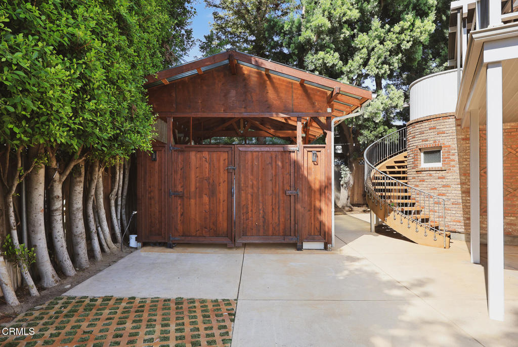 Wooden outbuilding with double doors beside a curved exterior staircase and brick wall.