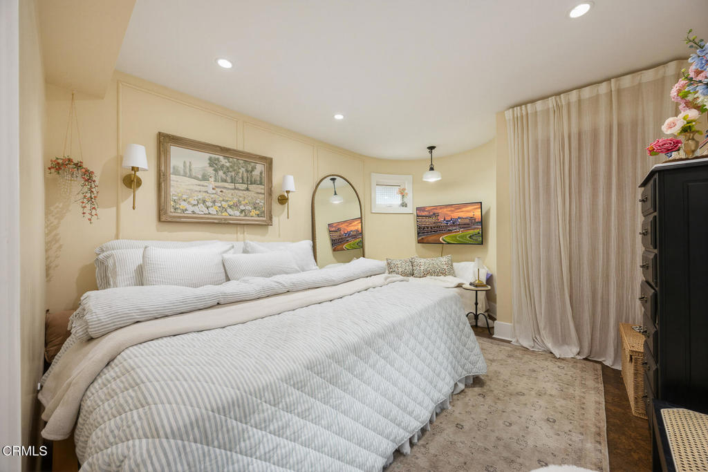 Bedroom with a large white bed, framed landscape art, arched mirror, and beige curtains.