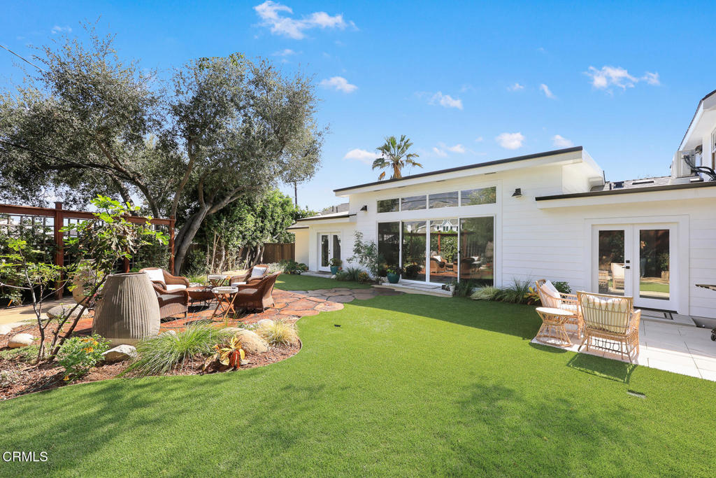 White single-story home with a broad lawn, landscaped patio seating, and large glass doors.