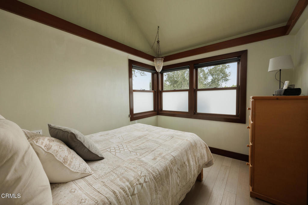 Bedroom with a patterned quilt, dark wood trim, and corner windows overlooking trees.
