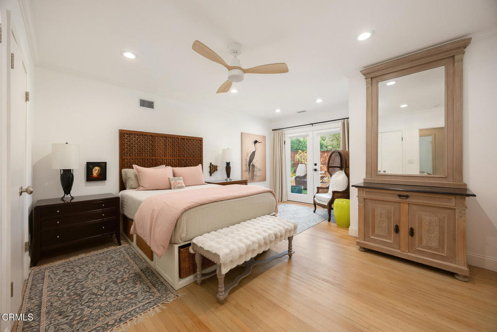 Furnished bedroom with blush bedding, carved wood headboard, tufted bench, and garden-facing glass doors.