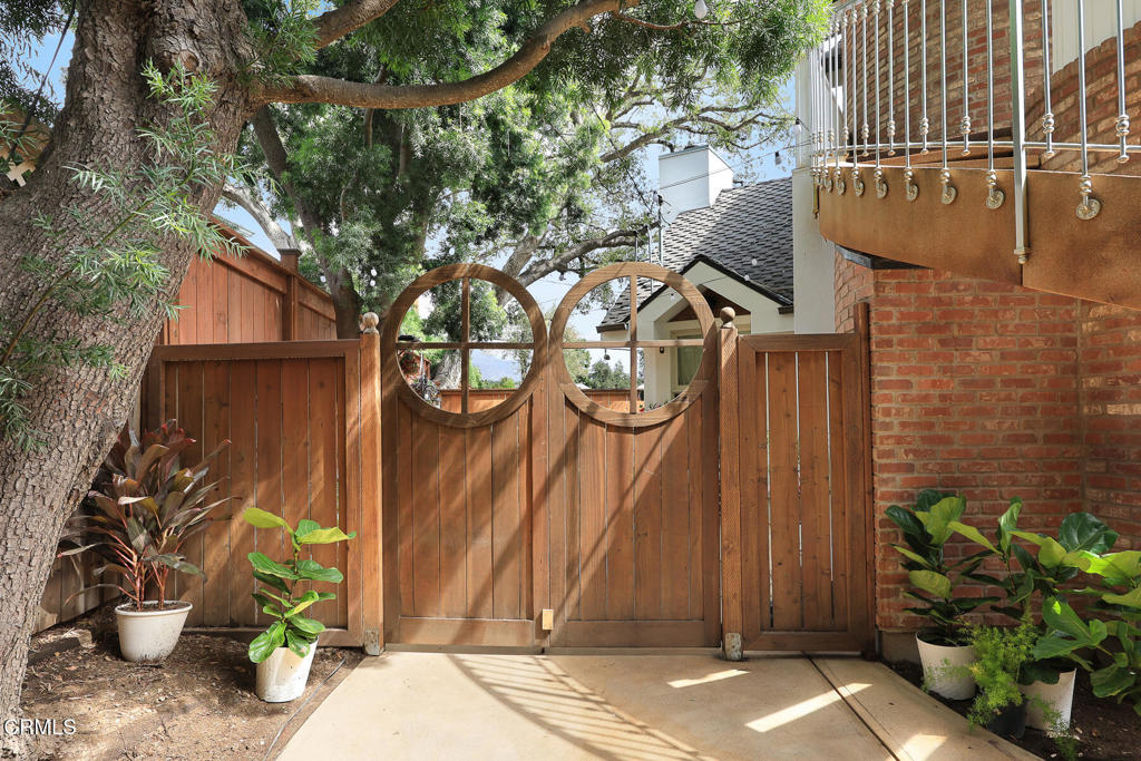 Wooden entry gate with circular cutouts beneath a large tree, beside a brick wall and balcony.