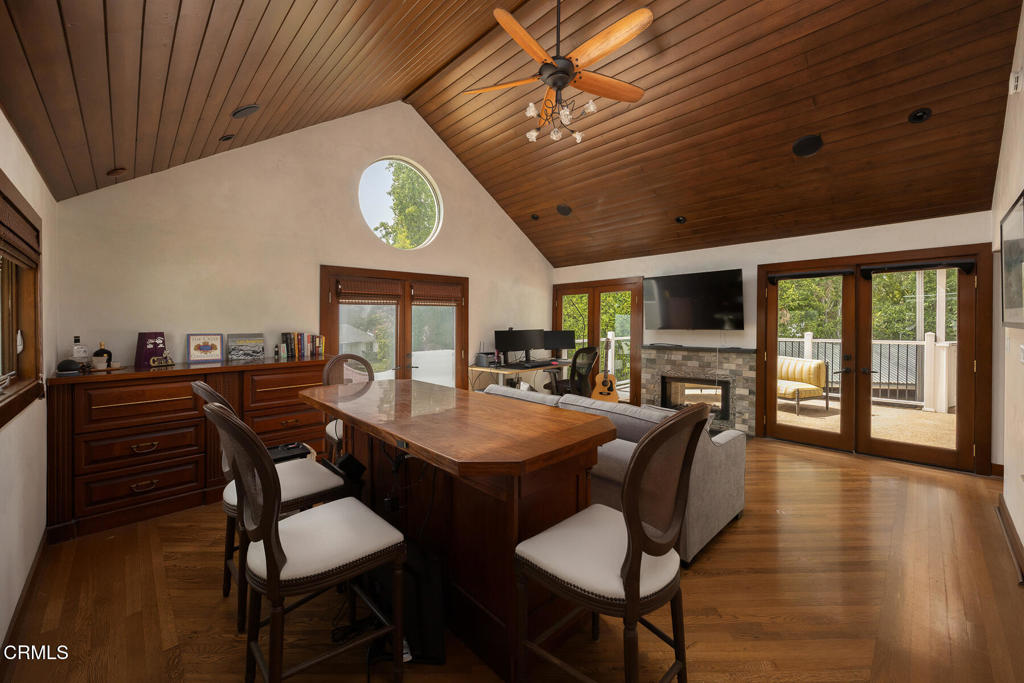 Dining and living area with a vaulted wood ceiling, large table, fireplace, and doors to a deck.