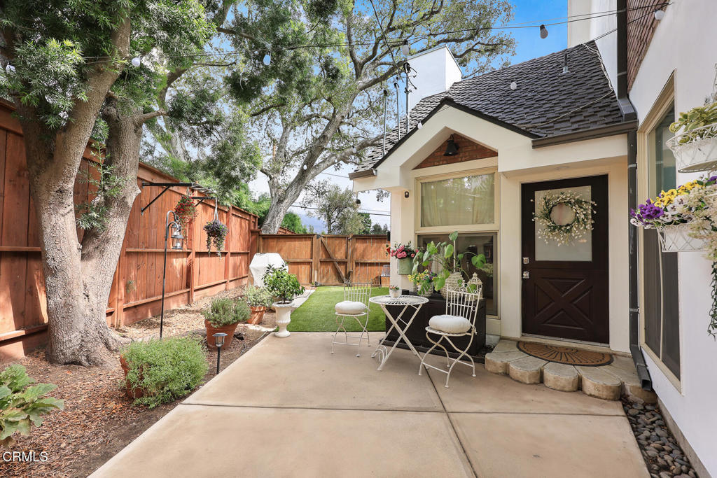 Backyard patio with white bistro seating, potted flowers, mature trees, and a redwood privacy fence.