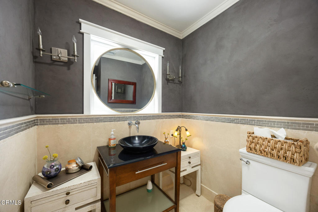 Bathroom with a round mirror, dark vessel sink, wood vanity, and woven basket atop the toilet.