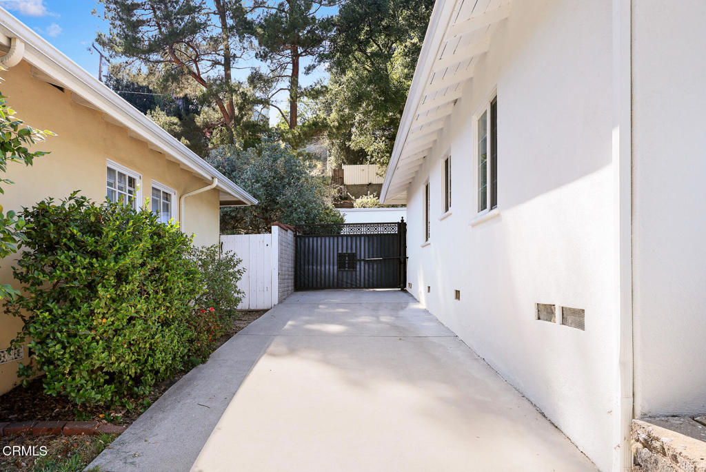 Concrete driveway leading to a black metal gate between two houses, bordered by shrubs and a white wall.