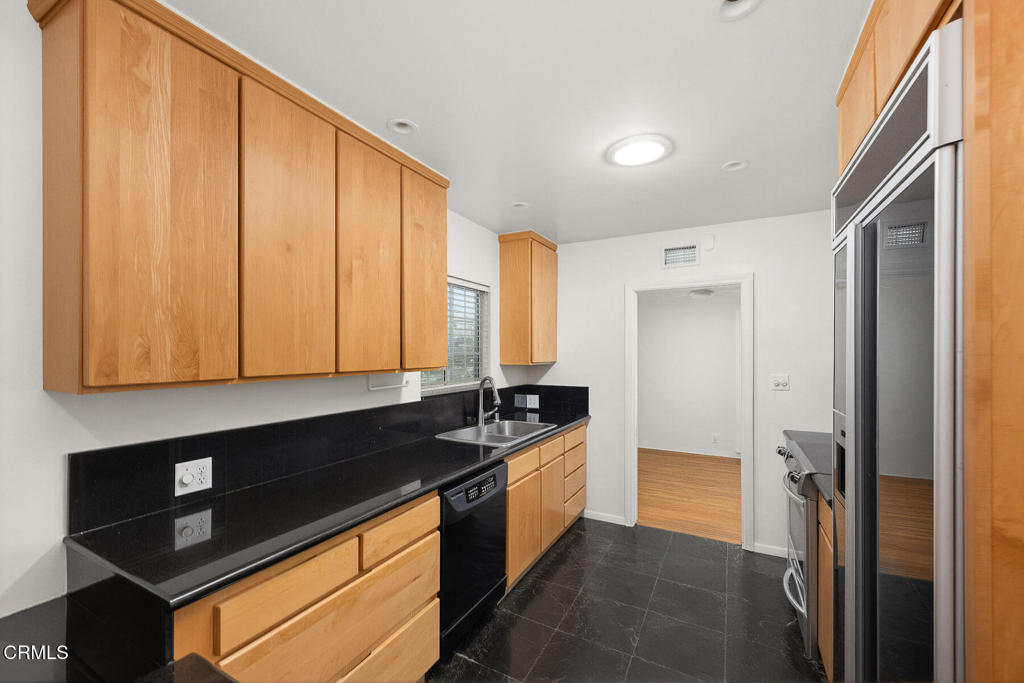 Galley kitchen with light wood cabinetry, black counters, dark floors, and built-in stainless-steel appliances.