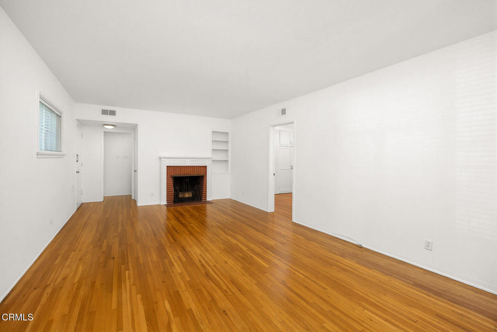 Vacant living room with hardwood floors, a brick fireplace, and built-in shelving.