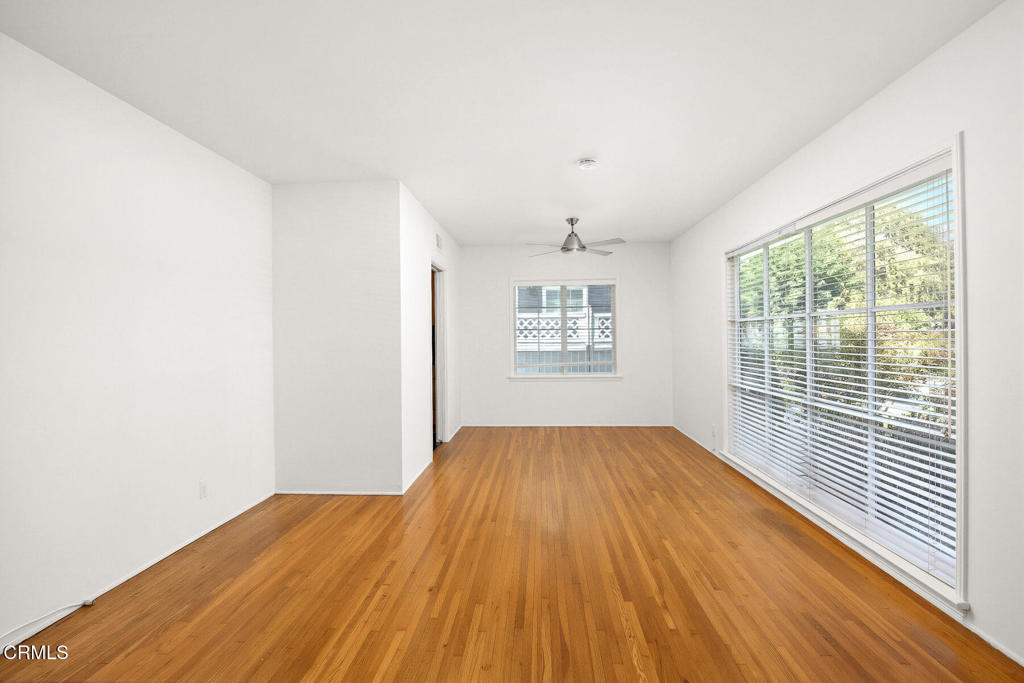 Empty room with warm hardwood floors, white walls, ceiling fan, and large window with blinds.