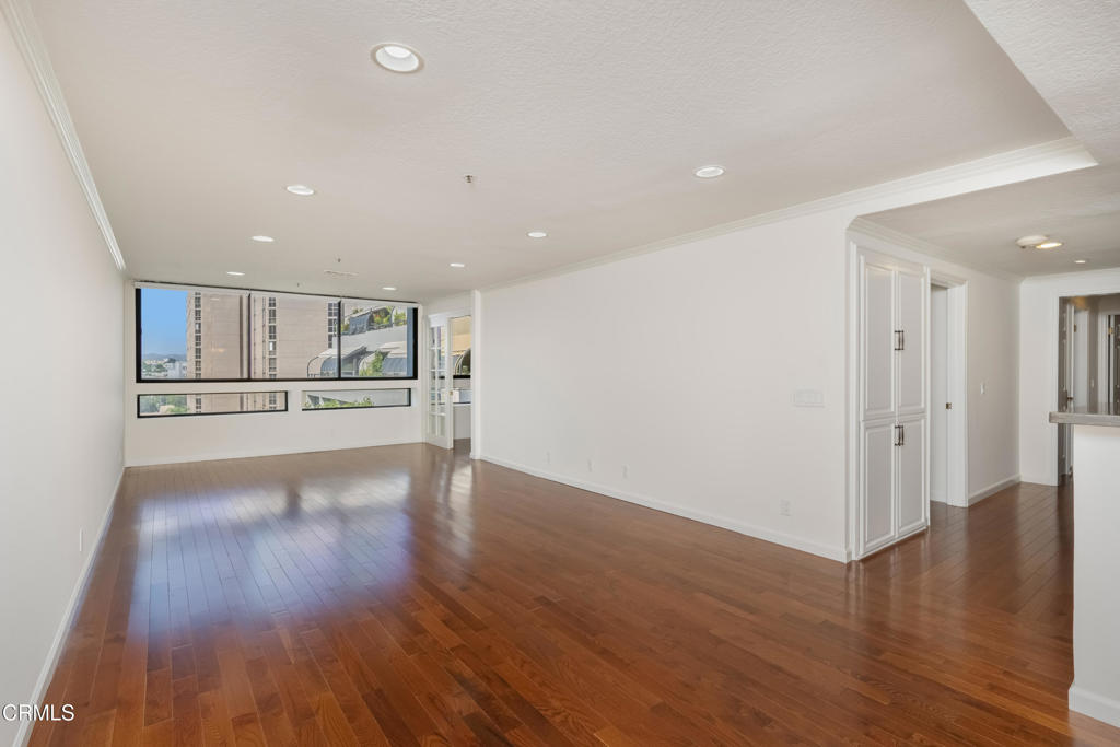 Empty living room with polished hardwood floors, wide windows, and built-in white cabinetry.
