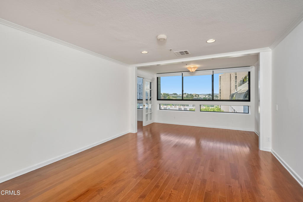 Vacant living area with hardwood floors and broad windows overlooking nearby buildings.