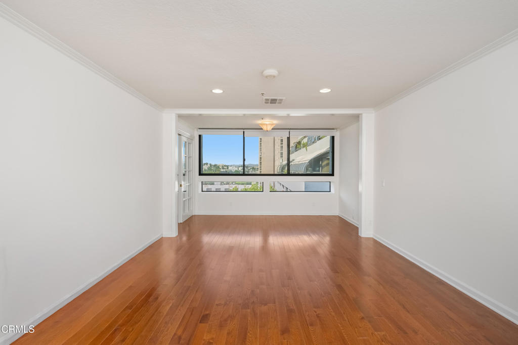 Vacant living room with polished wood floors and wide windows overlooking nearby buildings.