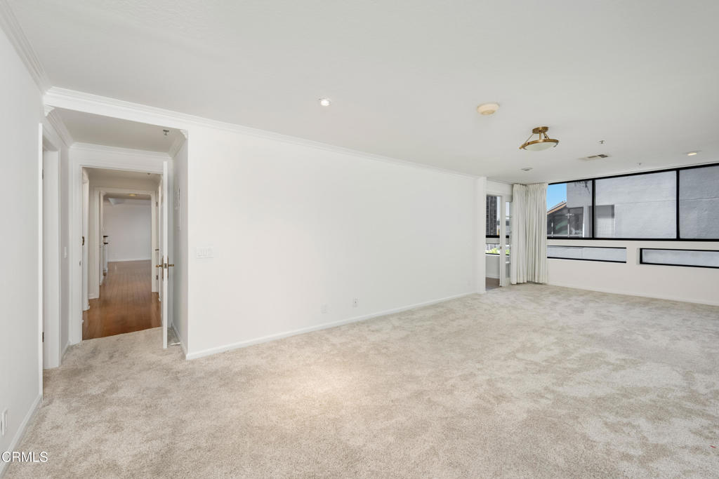 Empty carpeted living room with wide black-framed windows and a hallway leading off to the left.
