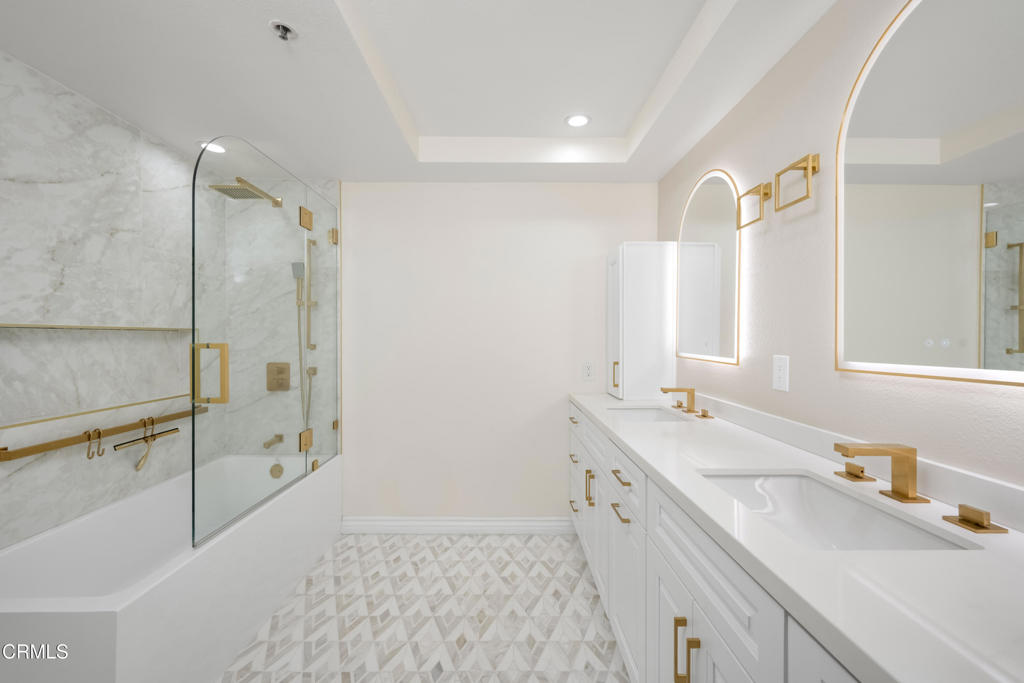 Bathroom with a double vanity, gold fixtures, glass-enclosed tub and shower, and geometric tile floor.