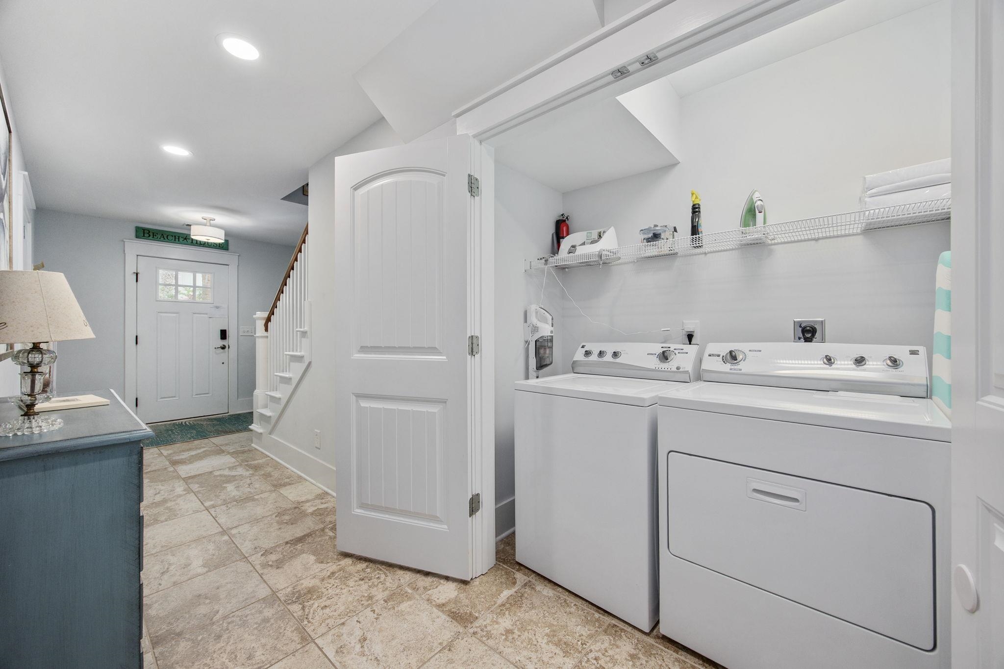 Laundry room with washer and dryer, wire shelving, tiled floor, and an open door to the entryway.