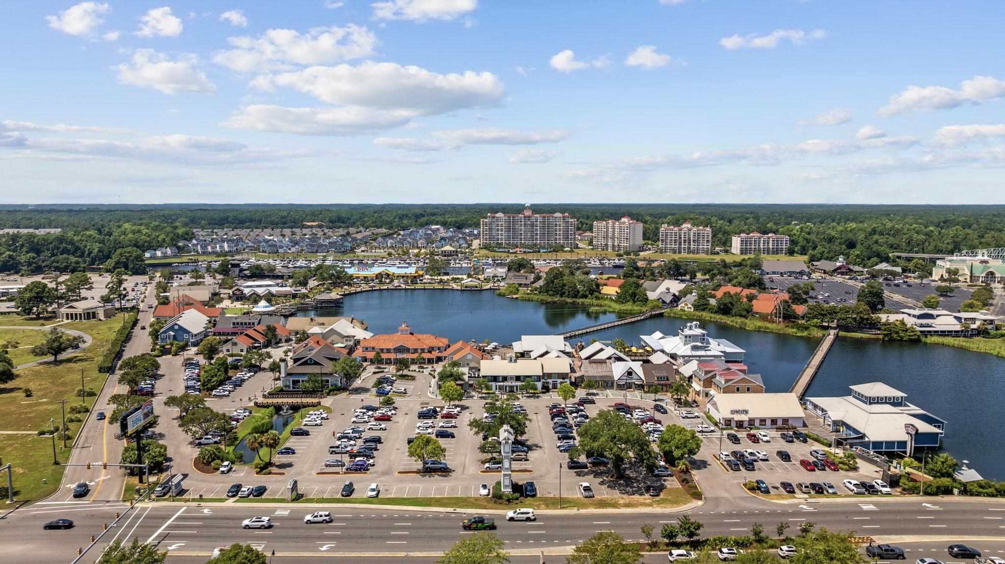 Aerial view of a waterfront shopping district with boardwalks, a pond, parking lots, and nearby condominiums.