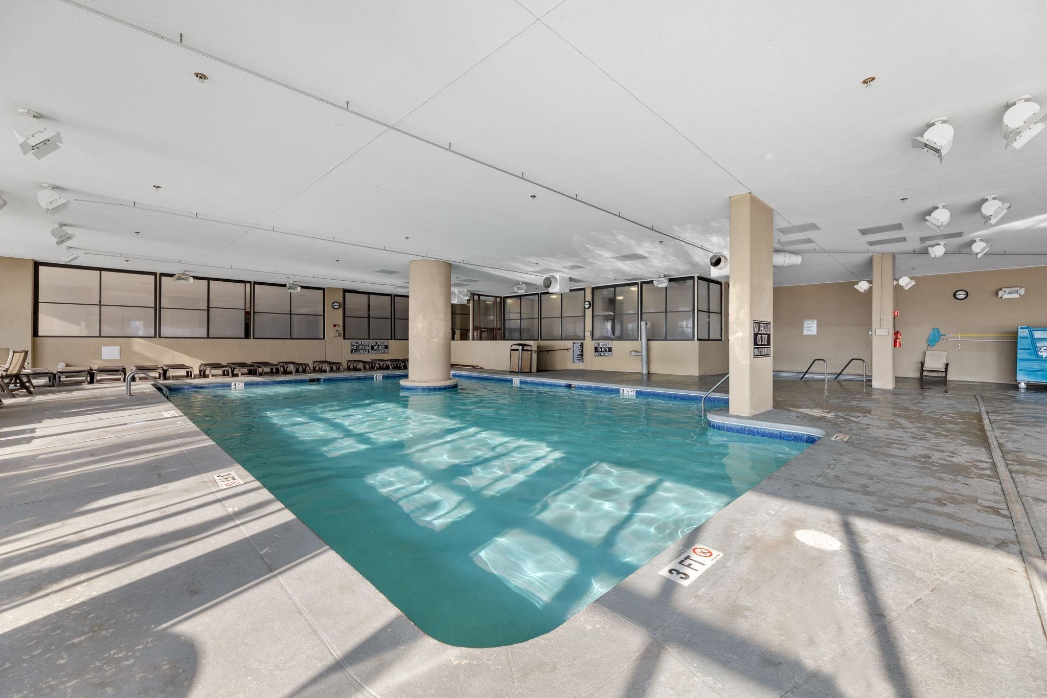 Indoor pool with turquoise water, a wide concrete deck, and lounge chairs along the windows.