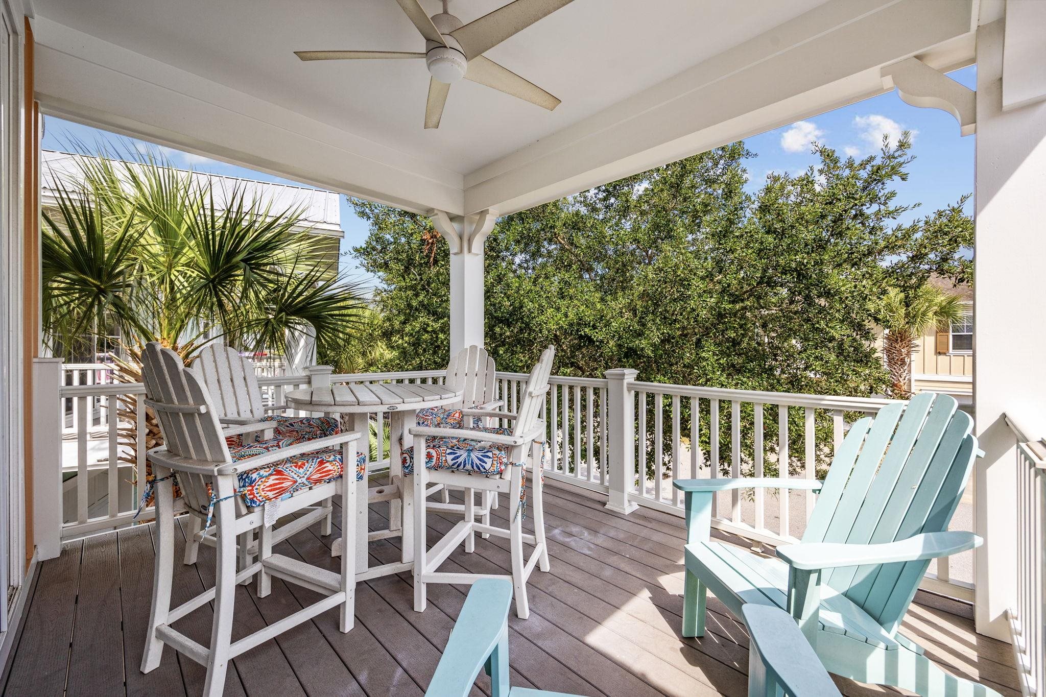 Covered balcony with white cushioned chairs, a round table, and turquoise Adirondack chairs.
