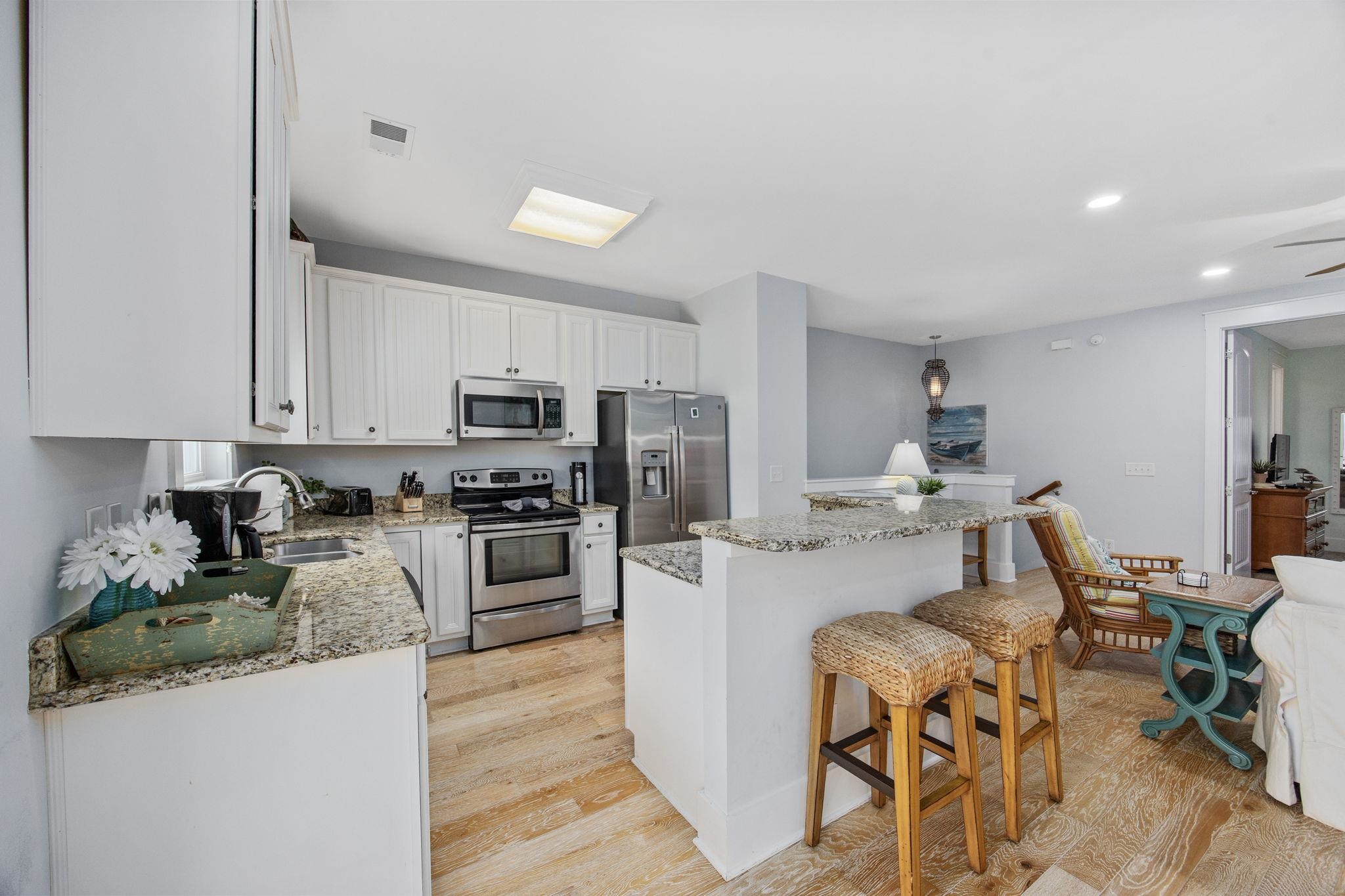 Open-plan kitchen with white cabinets, granite counters, stainless appliances, and a breakfast bar with woven stools.