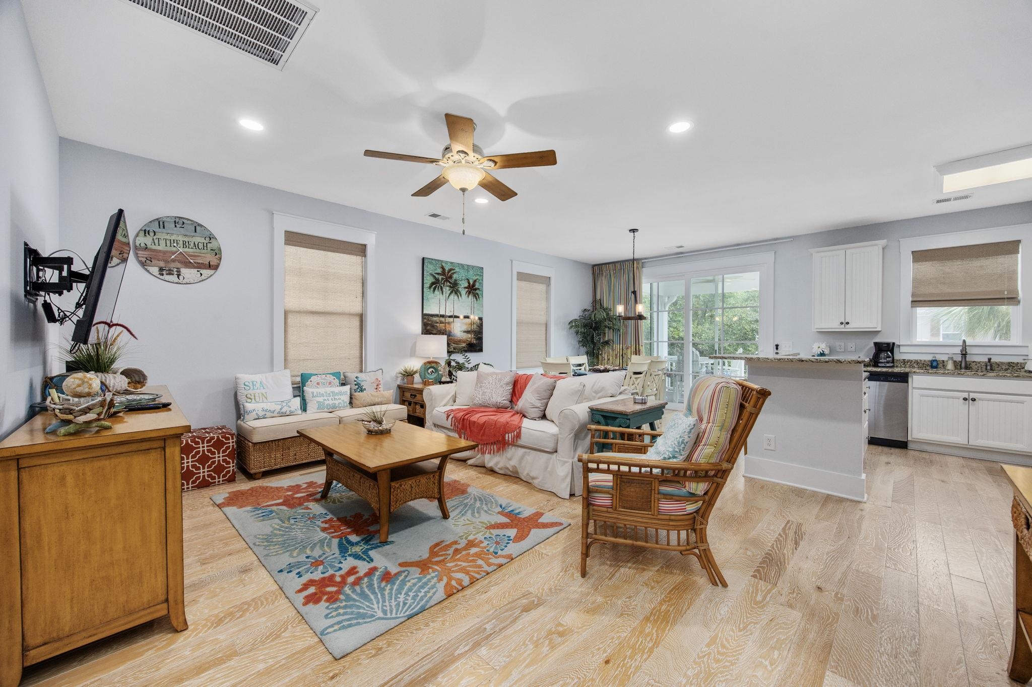 Furnished condo living room with white sofas, wicker chairs, a colorful shell-pattern rug, and balcony doors.