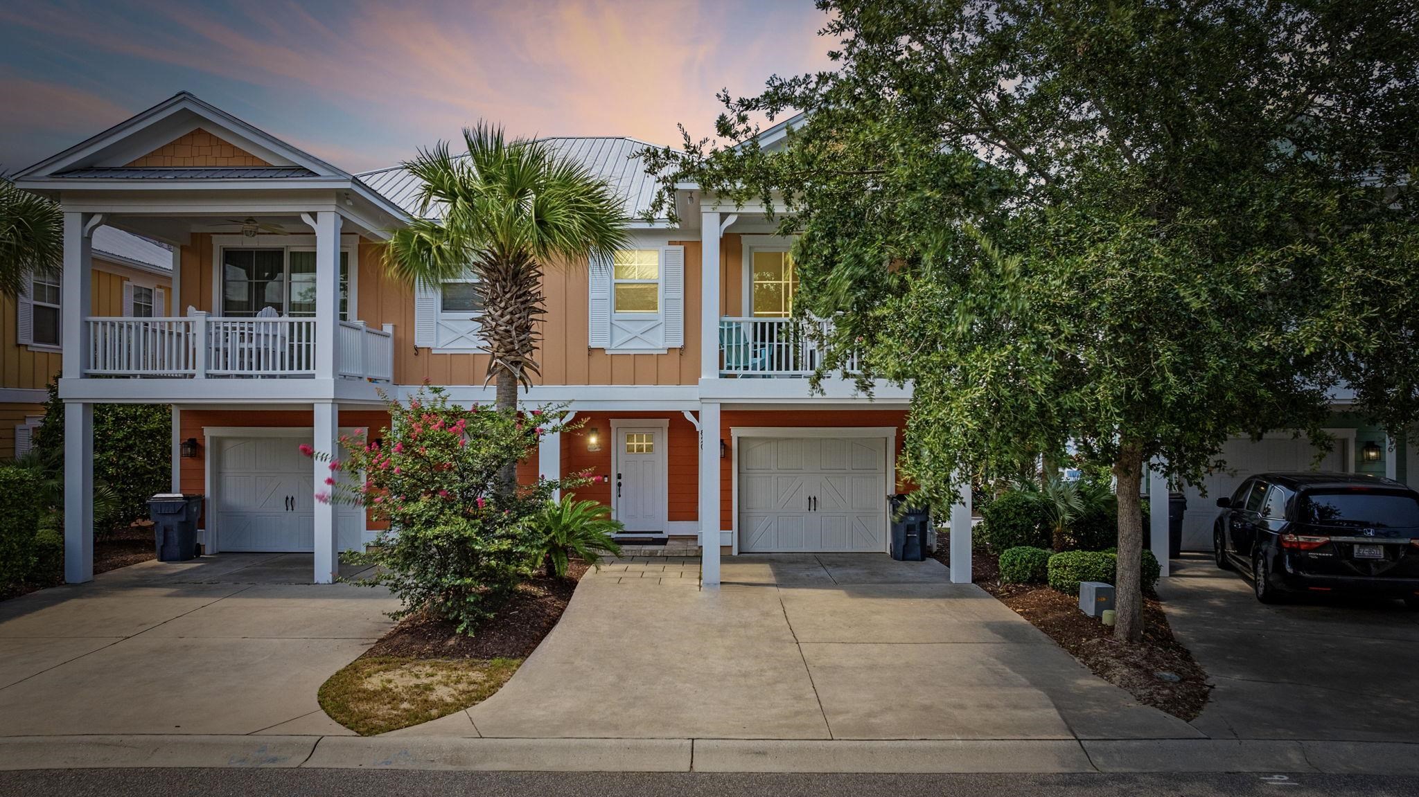 Two-story orange townhouse with white balconies and garage doors, framed by palms and flowering shrubs.