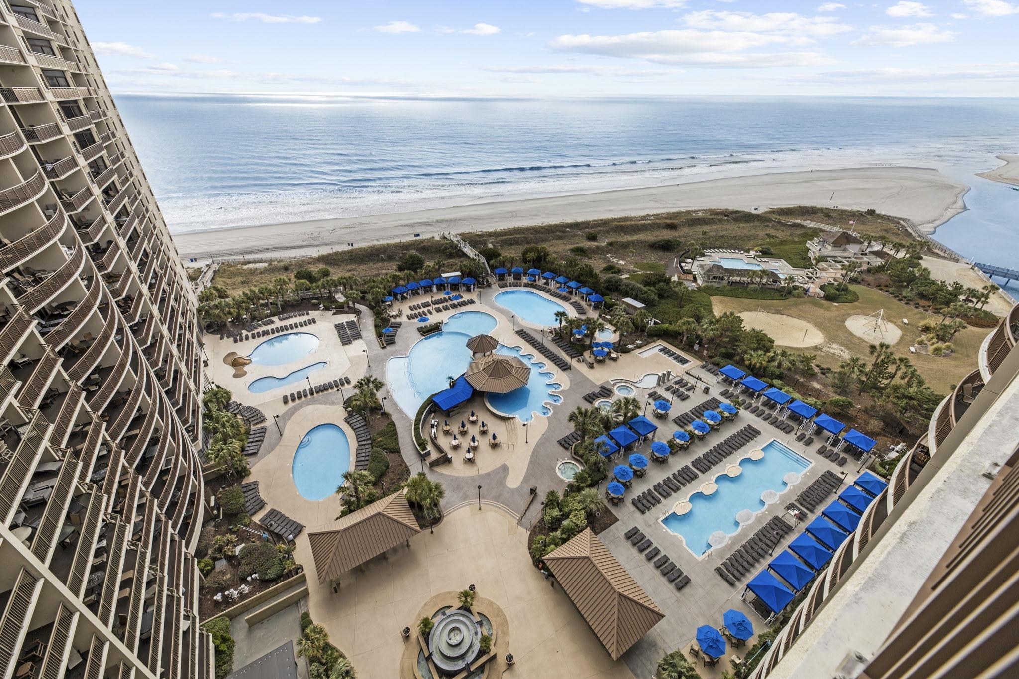 Aerial view of beachfront condo pools and blue-shaded lounge areas beside the beach and ocean.