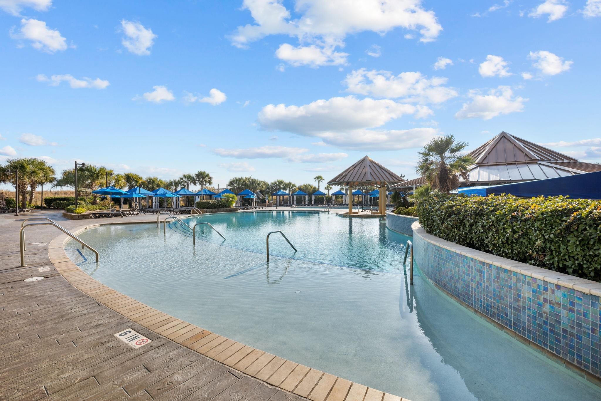 Community swimming pool with a curved shallow entry, blue umbrellas, and a palm-lined deck.