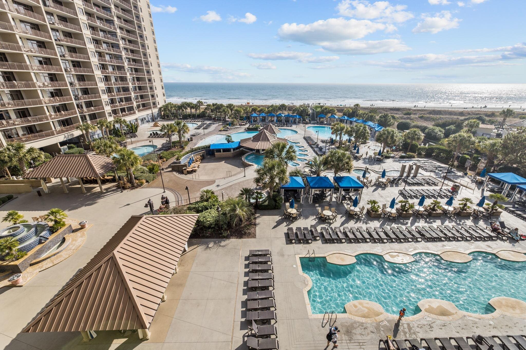Beachfront resort pools and cabanas beside a condominium tower, with the ocean beyond.