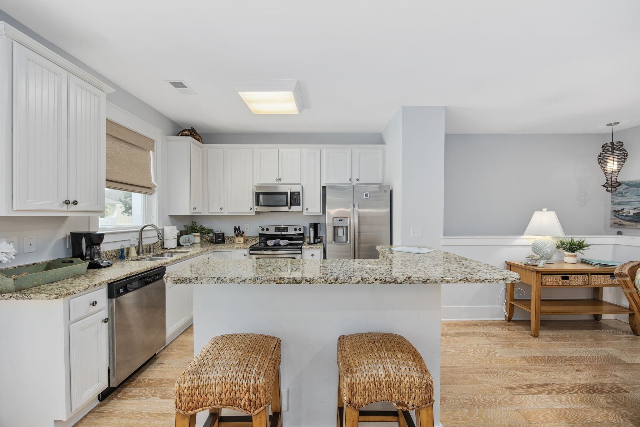 Kitchen with a large granite island, woven stools, white cabinets and stainless steel appliances.