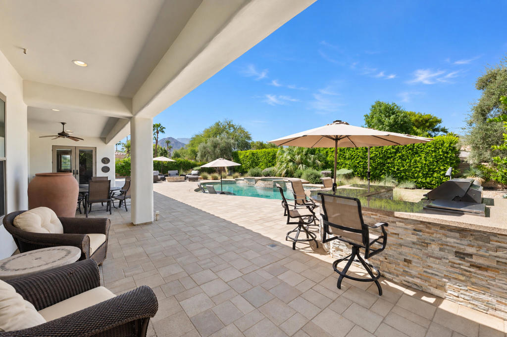 Covered patio overlooking a swimming pool, with lounge seating, umbrellas, and an outdoor grill.