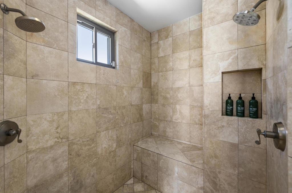 Walk-in shower with beige stone tile, a built-in bench, two showerheads, and a high window.