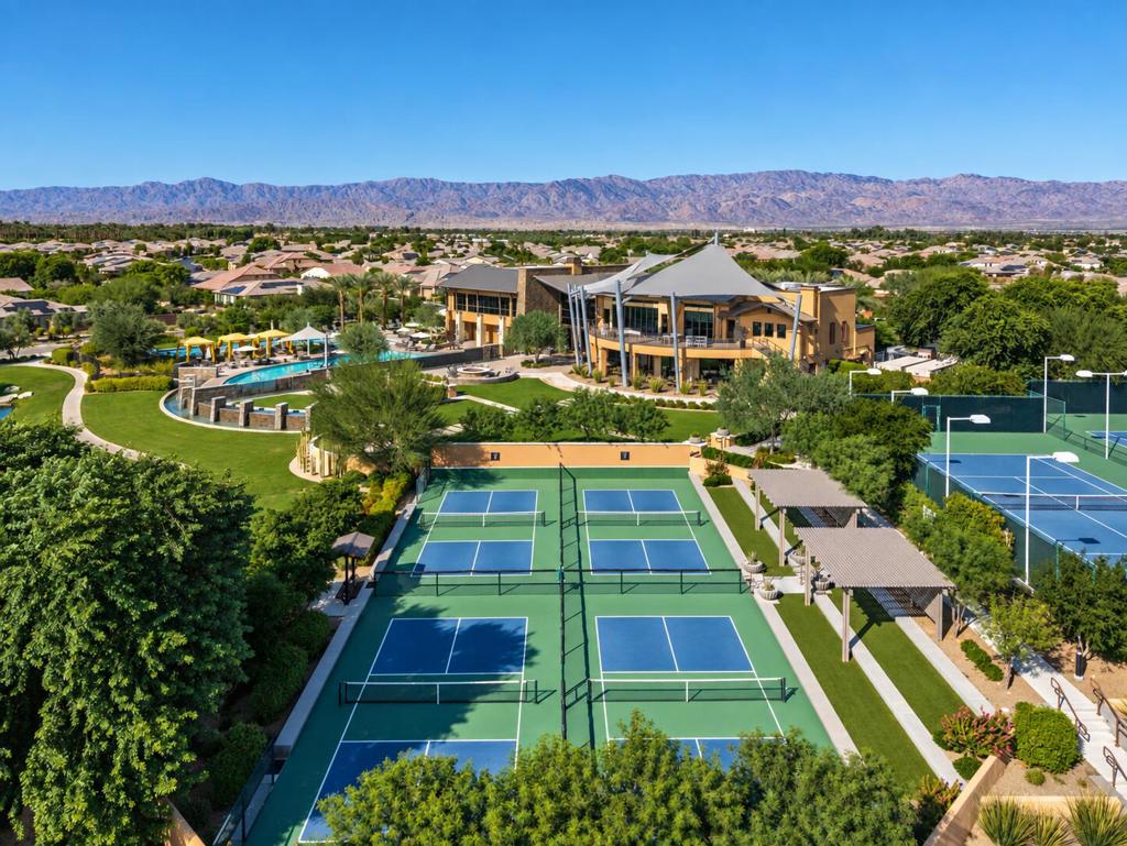 Aerial view of a desert community clubhouse, swimming pool, and tennis courts with mountains beyond.