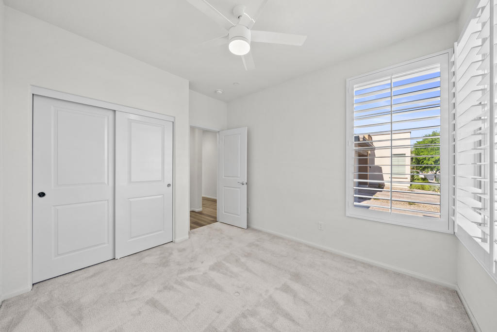Empty bedroom with light carpet, a ceiling fan, sliding closet doors, and shuttered windows.