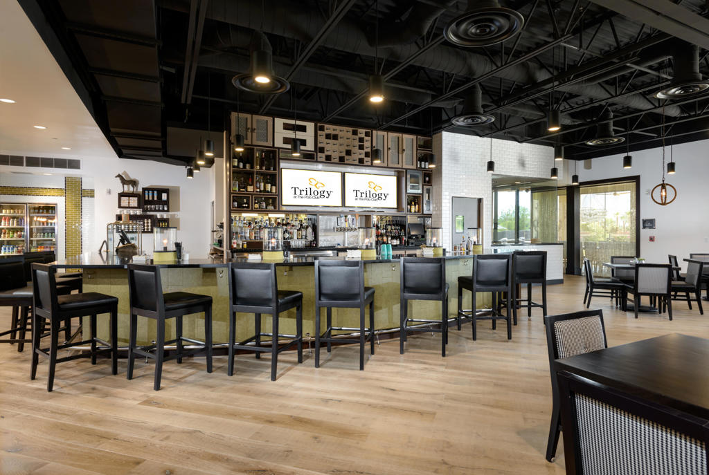 Clubhouse bar with a long counter, black barstools, bottle displays, and Trilogy signs.