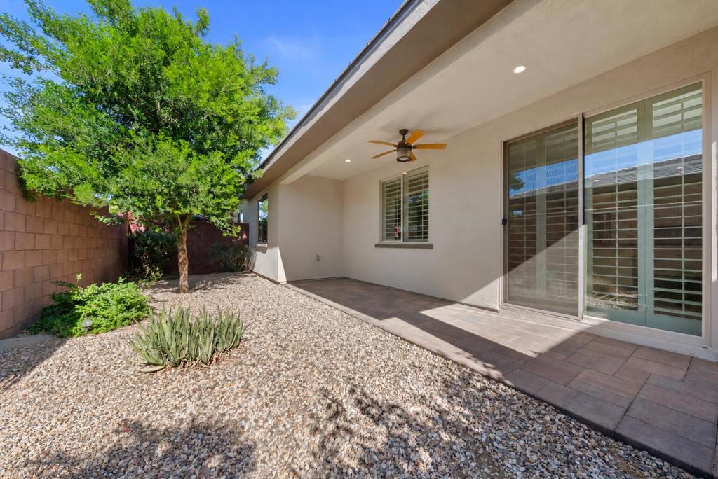Covered backyard patio with sliding glass doors, ceiling fan, gravel landscaping, and shade tree.