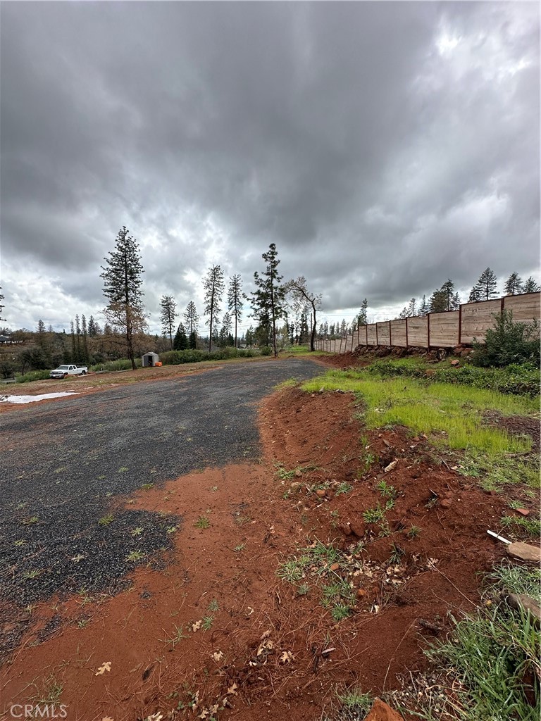 Gravel driveway bordered by red soil and a long wooden fence, with pine trees under cloudy skies.