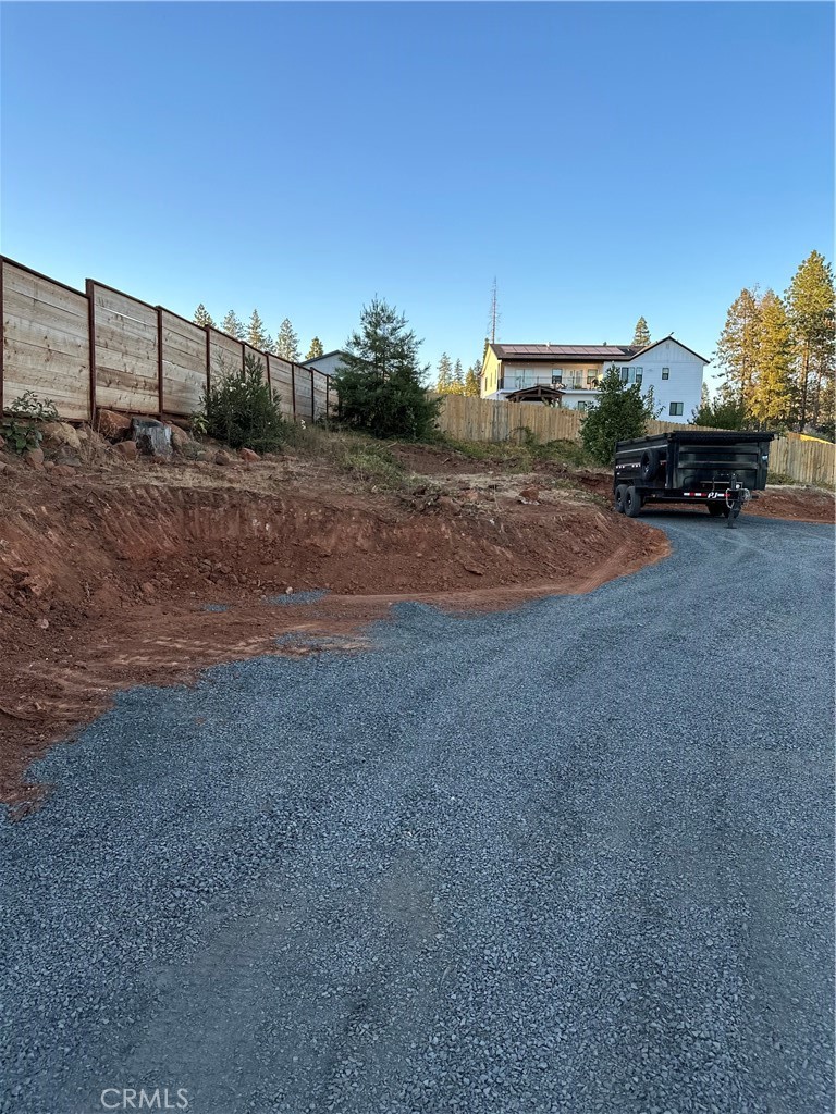 Gravel driveway curving toward a house, with graded soil, wooden fencing, and a black dump trailer.