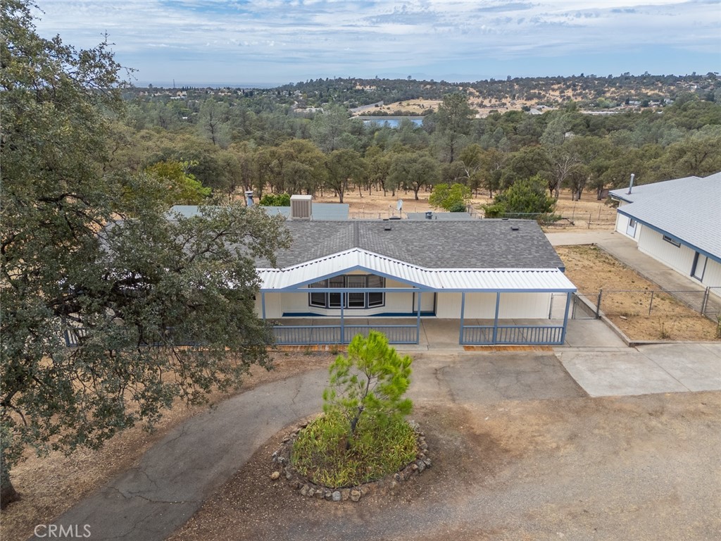 Aerial view of a single-story home with a covered porch, wooded surroundings, distant water, and hills.