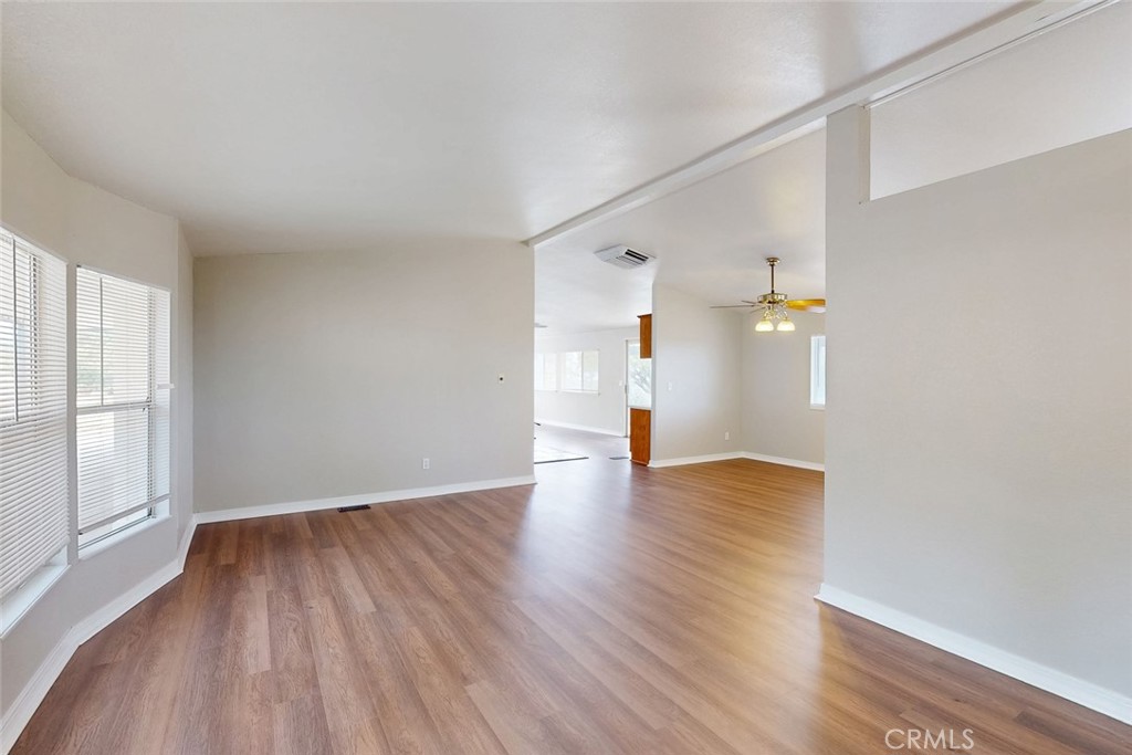 Vacant living and dining area with wood-look floors, window blinds, and an adjoining room with a ceiling fan.