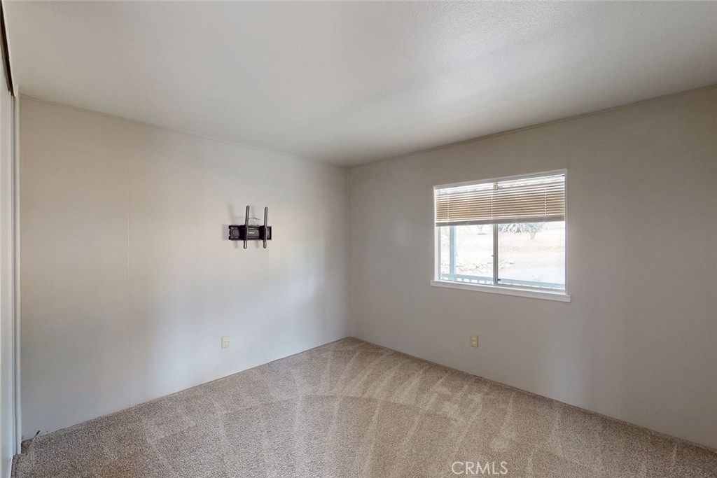 Vacant bedroom with beige carpet, a wall-mounted TV bracket, and a window with blinds.