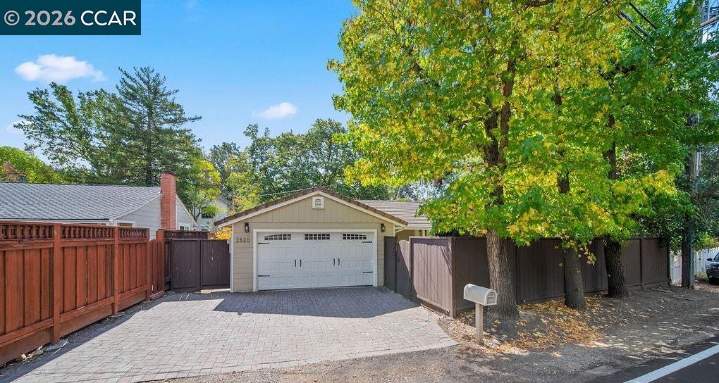 Paved driveway leading to a white garage, bordered by wooden fences and mature trees.
