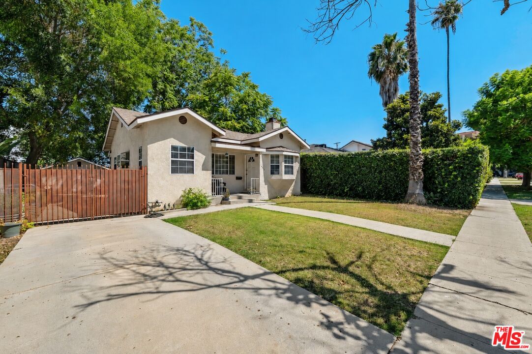 Beige stucco house with a wide concrete driveway, lawn, tall hedge, and palm trees.