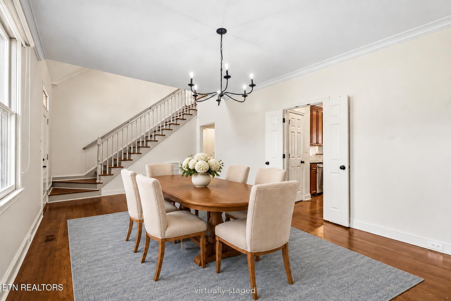 Dining room with a wooden table, upholstered chairs, black chandelier, and staircase.