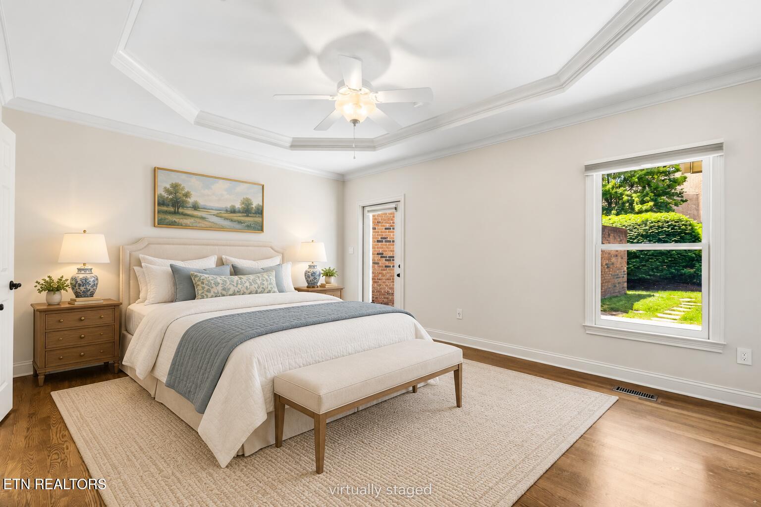 Staged bedroom with a tray ceiling, upholstered bed, and garden view through the window.